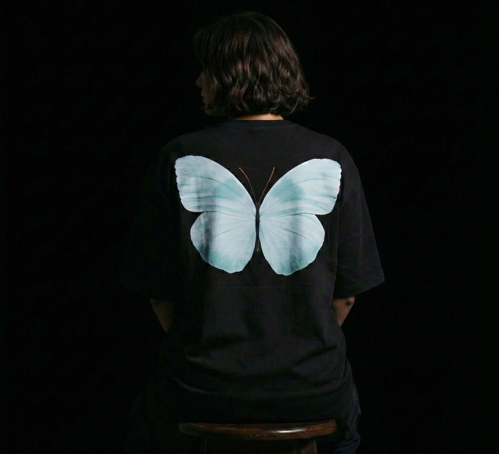 Person wearing a black t-shirt with a large blue butterfly design on the back against a dark background