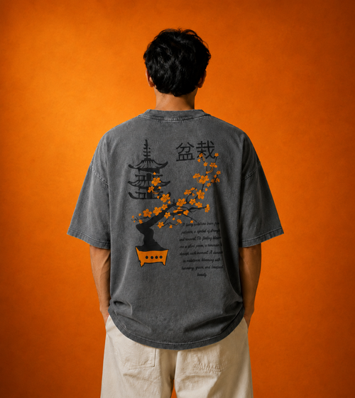 A man viewed from behind wearing a faded dark gray graphic t-shirt with a Japanese bonsai tree design and text, paired with light beige pants, set against a solid orange background.