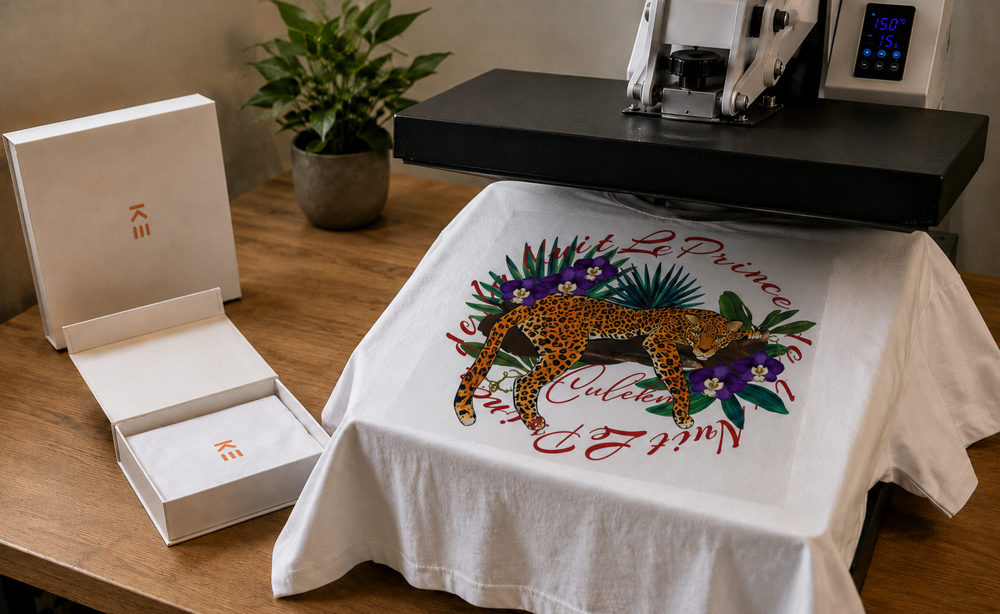 A white t-shirt with a colorful leopard graphic and red script text is being pressed flat under a heat press machine on a wooden table. An open white branded box with a gold logo sits to the left, with a small potted plant in the background.