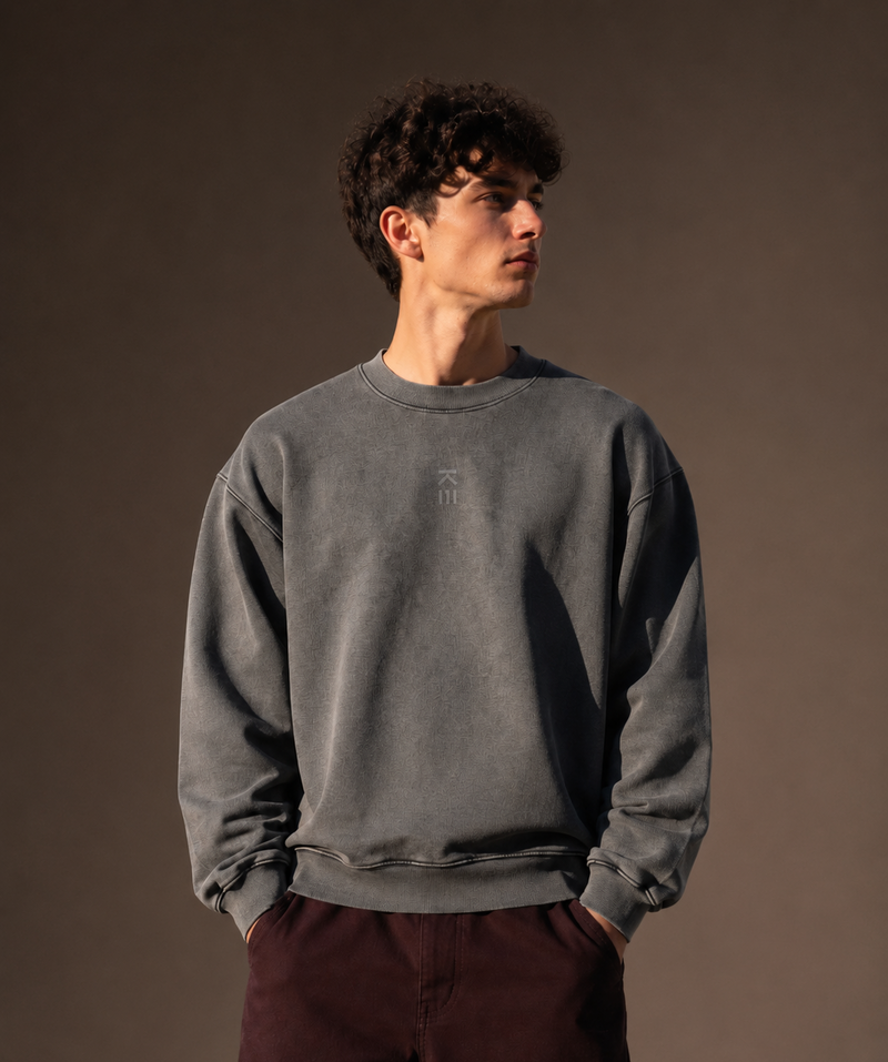 A man wearing a washed dark gray crewneck sweatshirt and dark burgundy pants, standing against a solid dark brown studio background.