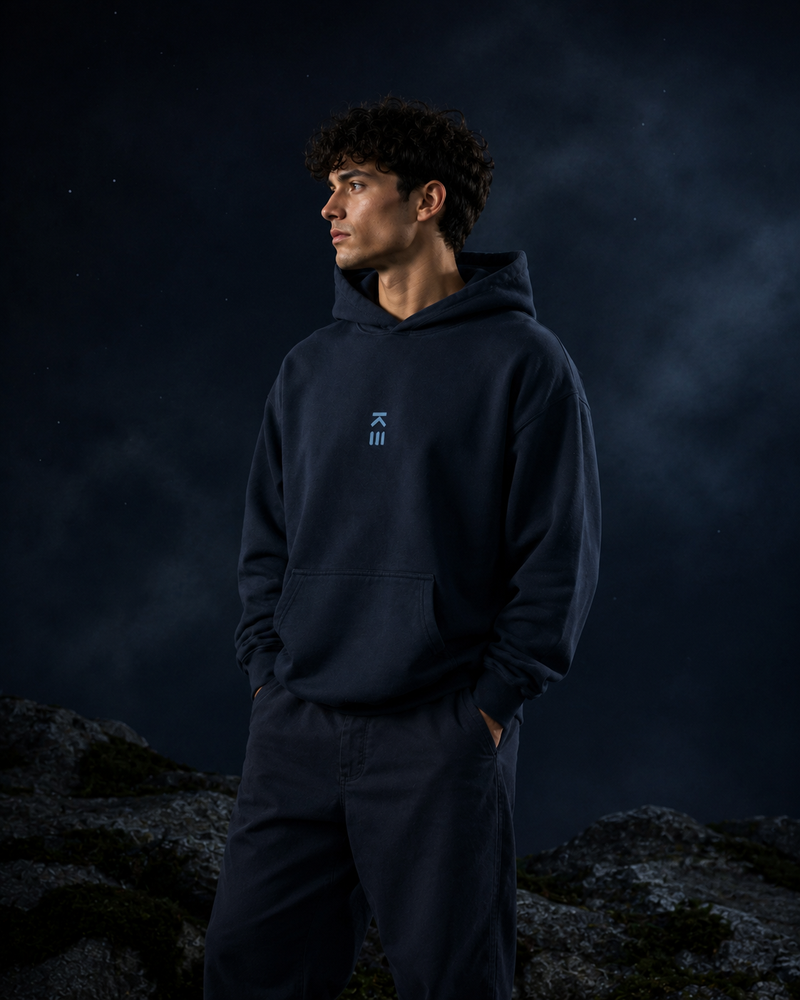 A man wearing a dark navy blue hoodie and matching sweatpants stands outdoors against a dark, moody night sky with faint clouds and rocky ground visible.