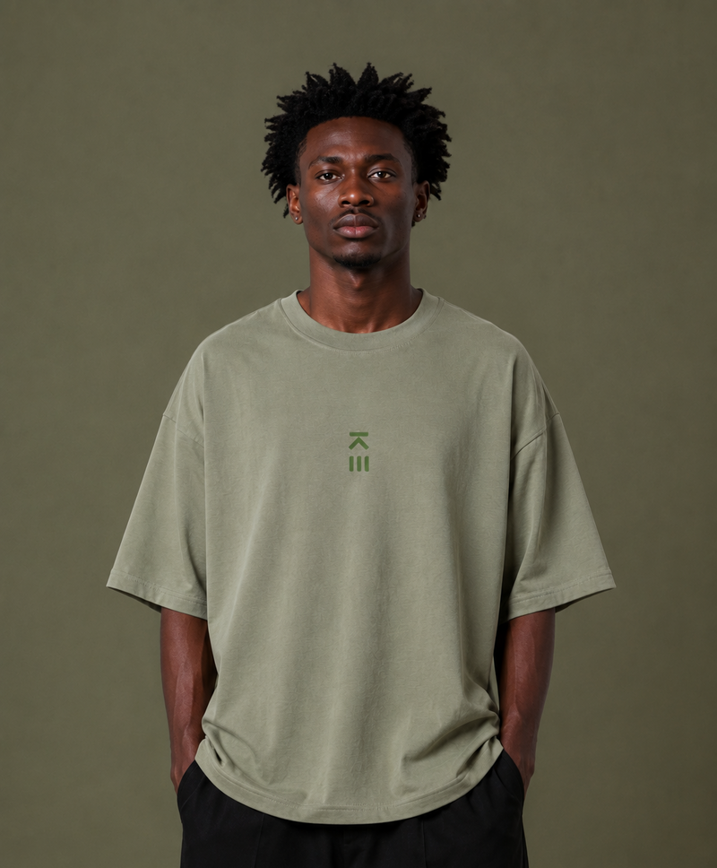 A man wearing an oversized sage green t-shirt with a small green logo on the chest, paired with black pants, standing against a solid olive green background.